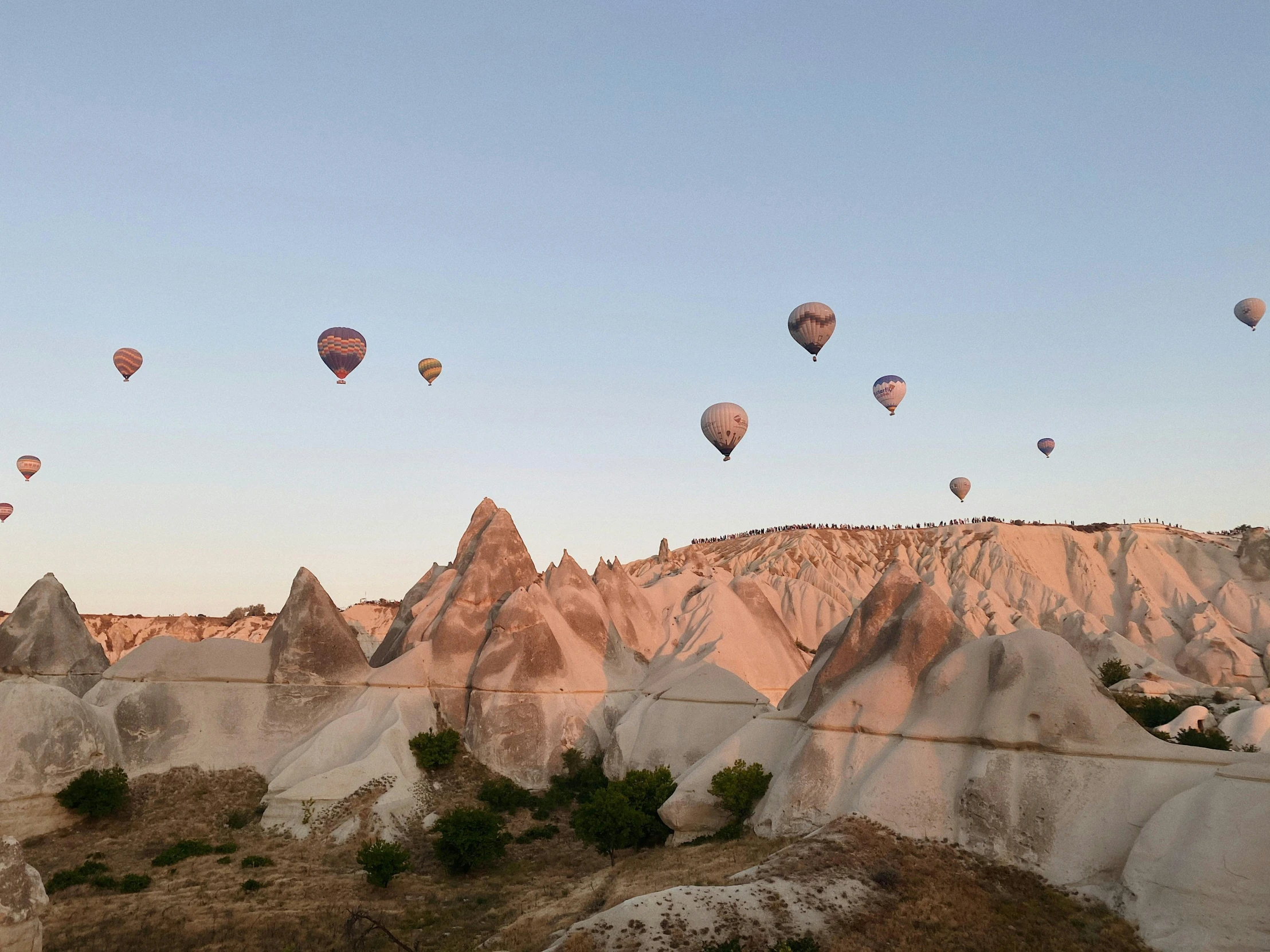 a group of hot air balloons flying over a valley, a photo, pexels contest winner, art nouveau, white travertine terraces, cliff side at dusk, grayish, blue sky