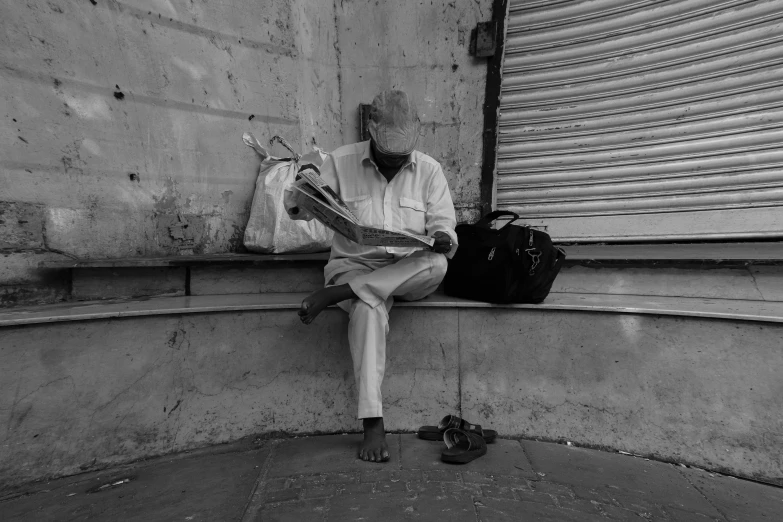 a man sitting on a ledge reading a newspaper, a black and white photo, streets of mumbai, baggy clothing and hat, trending photo, an oldman