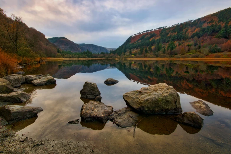 a body of water surrounded by rocks and trees, by Bedwyr Williams, pexels contest winner, irish mountains background, floating stones, colors reflecting on lake, thumbnail