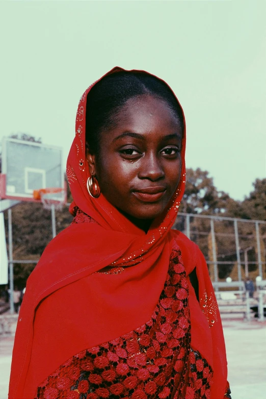 a woman in a red shawl stands in front of a basketball court, inspired by Ras Akyem, pexels contest winner, black teenage girl, halfbody headshot, color film still, wearing an african dress