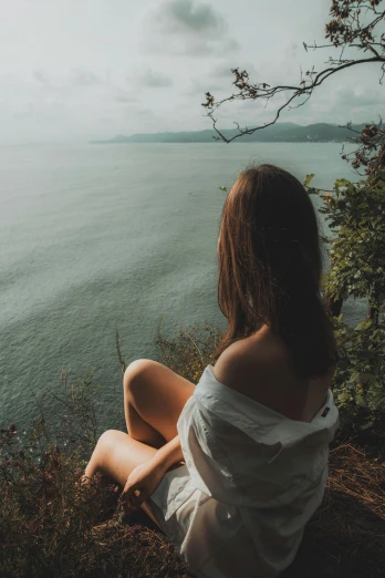 a woman sitting on the edge of a cliff overlooking a body of water, a picture, inspired by Elsa Bleda, trending on unsplash, renaissance, bare shoulders, panoramic view of girl, lush surroundings, view of the ocean