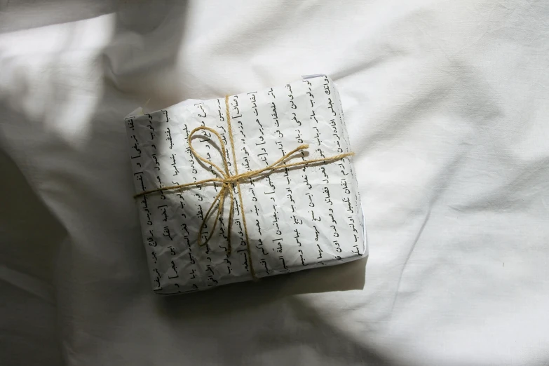 a wrapped gift sitting on top of a bed, by Alice Mason, unsplash, hurufiyya, calligraphic poetry, white splendid fabric, background image, ignant