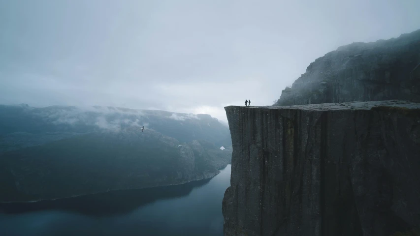 a person standing on the edge of a cliff, by Anton Lehmden, pexels contest winner, realism, grey mist, tiny people walking below, nordic, 8k resolution”