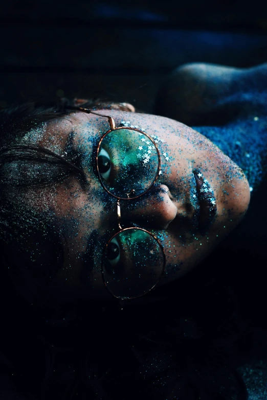 a woman laying on top of a bed covered in blue paint, inspired by Elsa Bleda, trending on pexels, wearing gold glasses, dark glitter, face in water, galactic dark colors