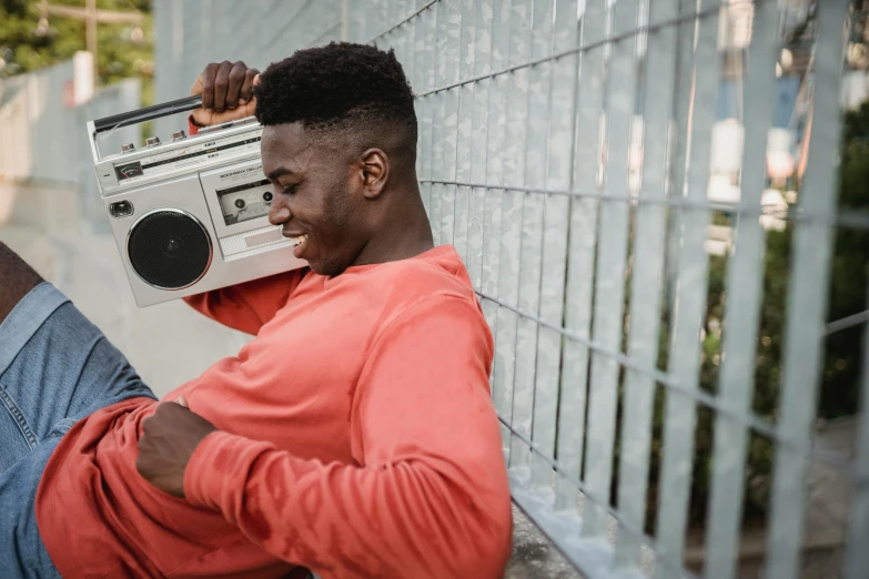 a man leaning against a fence holding a boombox, pexels contest winner, happening, black teenage boy, music being played, laughing, slightly muscular