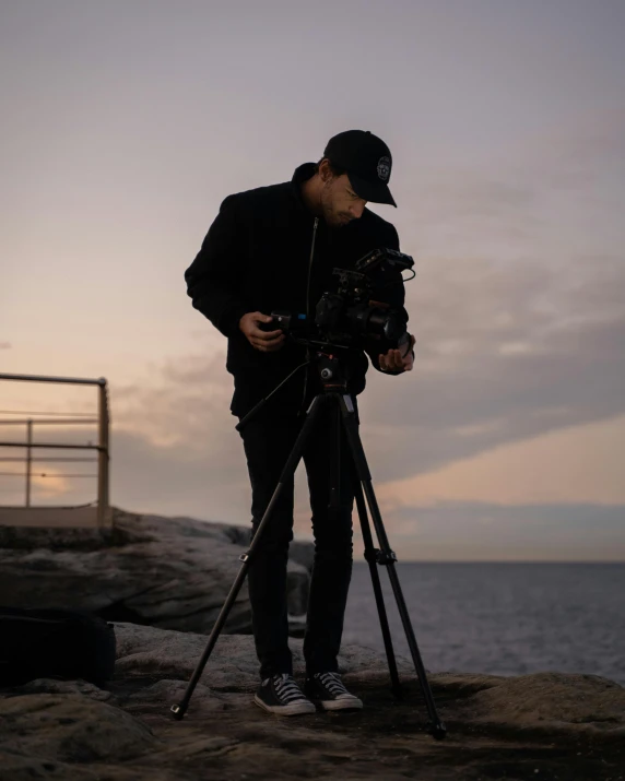 a man standing on top of a beach next to a camera, pexels contest winner, video art, cinematic outdoor lighting, movies, a man wearing a black jacket, home video footage