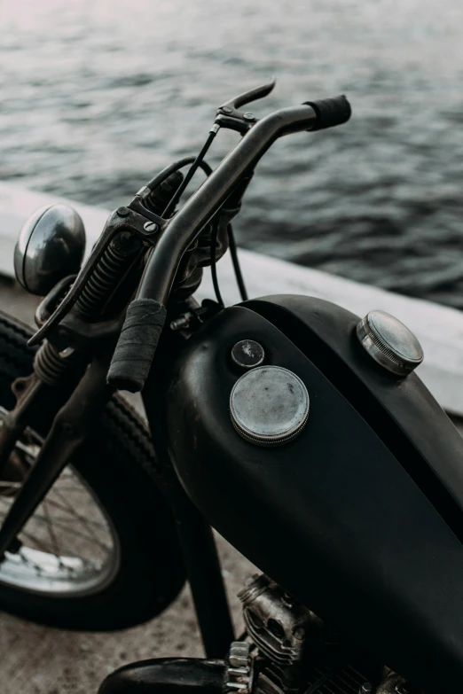 a black motorcycle parked next to a body of water, pexels contest winner, modernism, mechanical detail, 1952, grey, bumpy skin