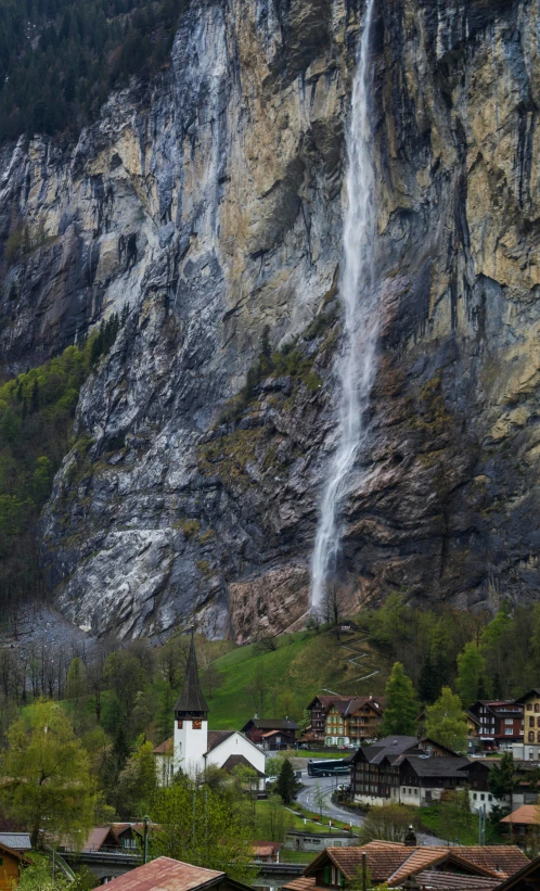 a waterfall is coming down the side of a mountain, by Franz Hegi, lauterbrunnen valley, behind a tiny village, panorama shot, taken in the late 2010s