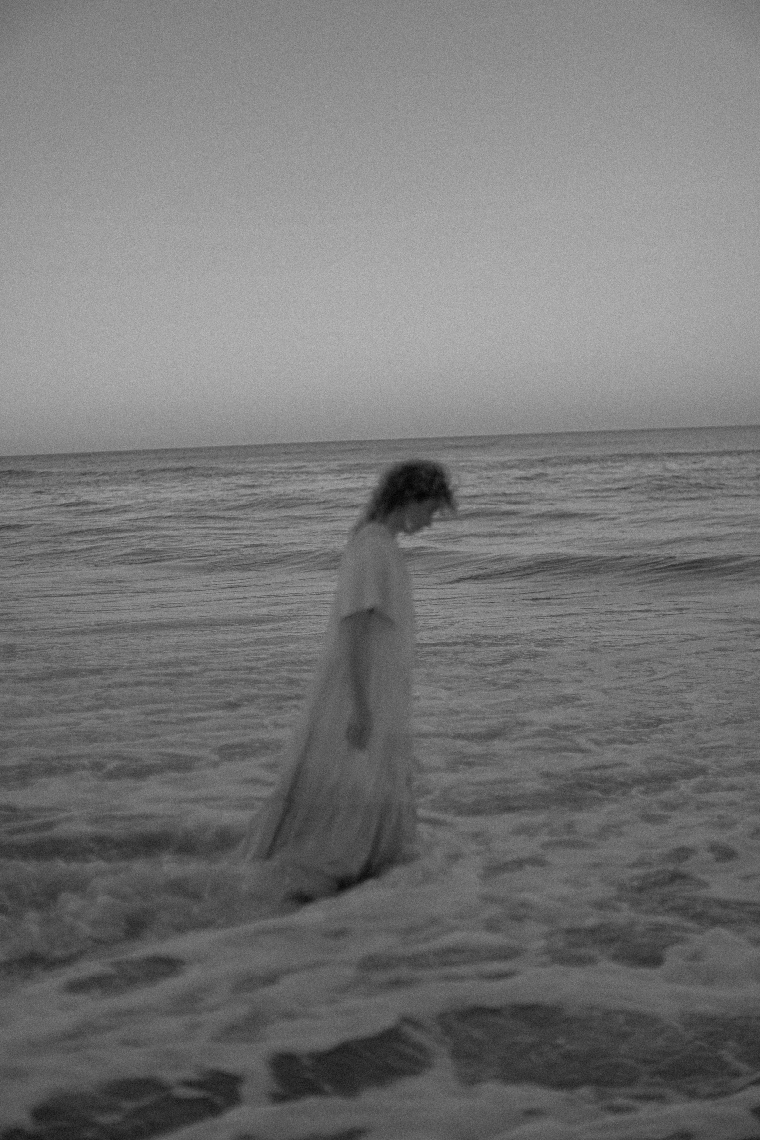 a woman standing on top of a beach next to the ocean, a black and white photo, tumblr, pale young ghost girl, alternate album cover, ffffound, dusk