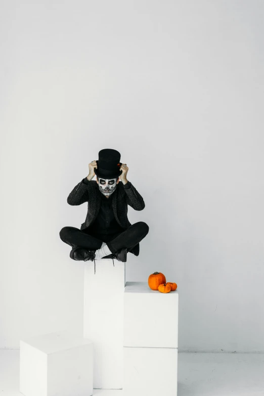 a man sitting on top of a white cube, pexels contest winner, vanitas, in a halloween style, white backdrop, black hat, hear no evil