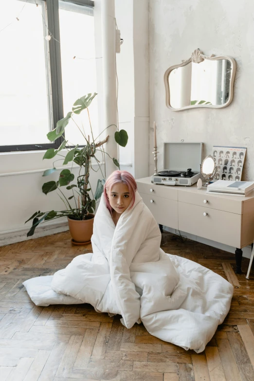 a woman sitting on a floor covered in a blanket, an album cover, inspired by Elsa Bleda, unsplash, with pink hair, wearing white robe, standing in corner of room, model wears a puffer jacket