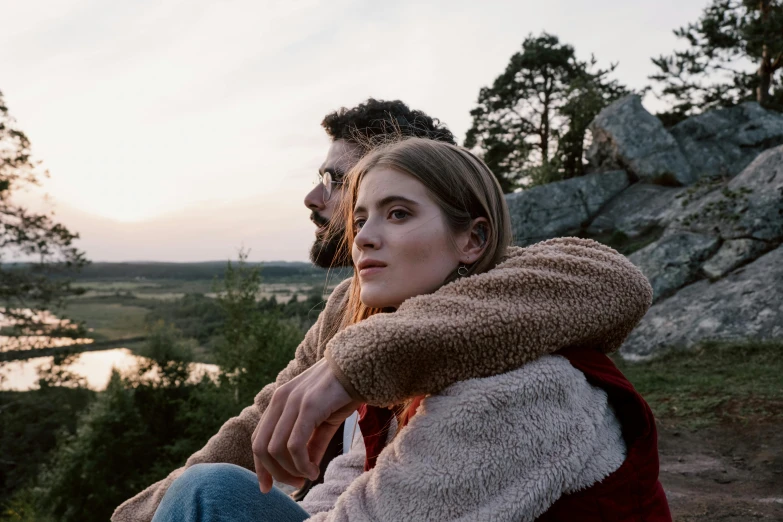 a man and a woman sitting next to each other, a picture, by Adam Marczyński, trending on pexels, romanticism, looking off into the distance, still from the movie, arm around her neck, cliffside