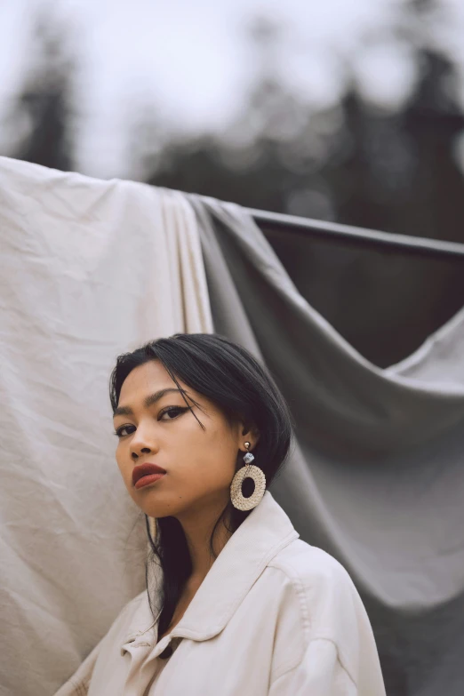 a woman standing in front of a white sheet, an album cover, inspired by Ruth Jên, unsplash, earrings, south east asian with round face, outdoor photo, ((portrait))