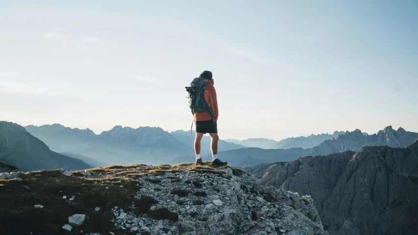 a man standing on top of a mountain with a backpack, by Niko Henrichon, pexels contest winner, al fresco, profile image, white shorts and hiking boots, facing sideways