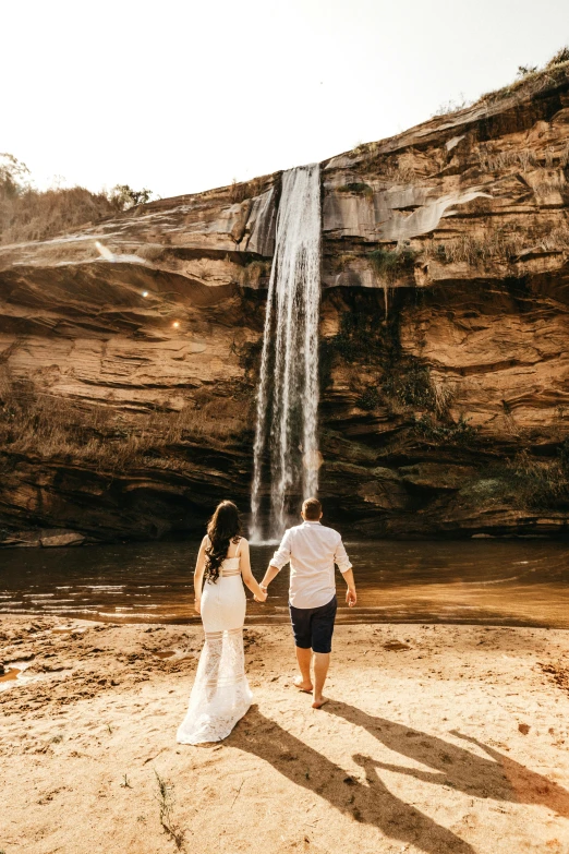 a bride and groom standing in front of a waterfall, by Lee Loughridge, unsplash contest winner, walking on the sand, waterfall below, back, tourist photo