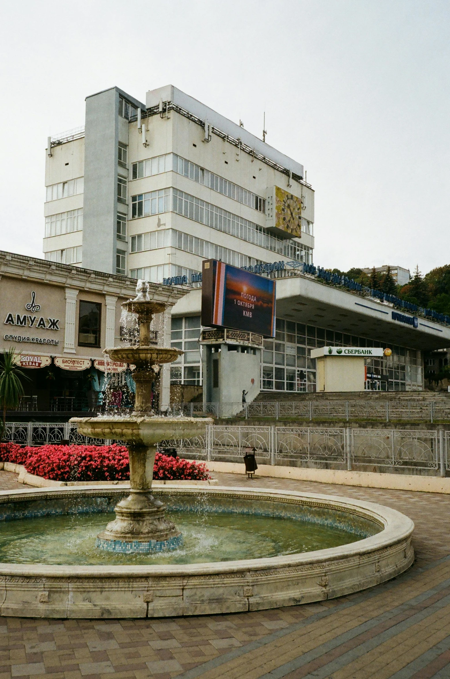 a fountain in a plaza with a building in the background, inspired by Thomas Struth, art nouveau, soviet town, hillside, cinema, lots of shops