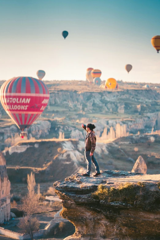 a man standing on the edge of a cliff with hot air balloons in the background, taking a picture, silvain sarrailh, color photo, killian eng