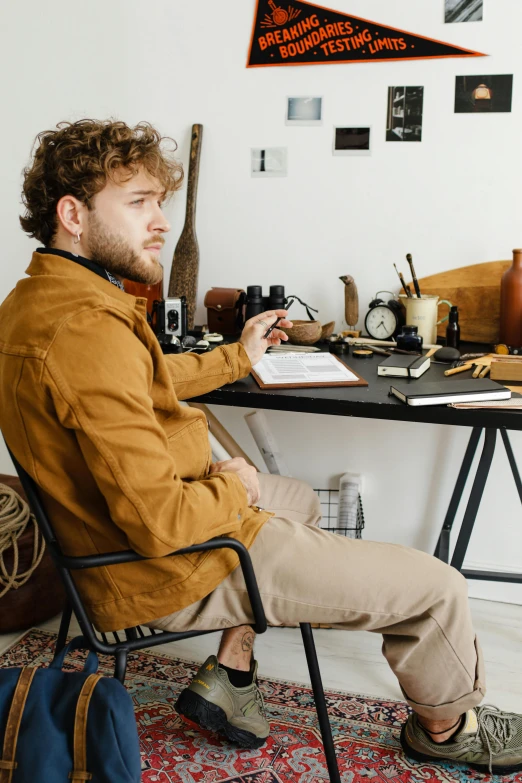a man sitting in a chair in front of a desk, trending on pexels, arbeitsrat für kunst, brown clothes, curly copper colored hair, studious, starving artist wearing overalls
