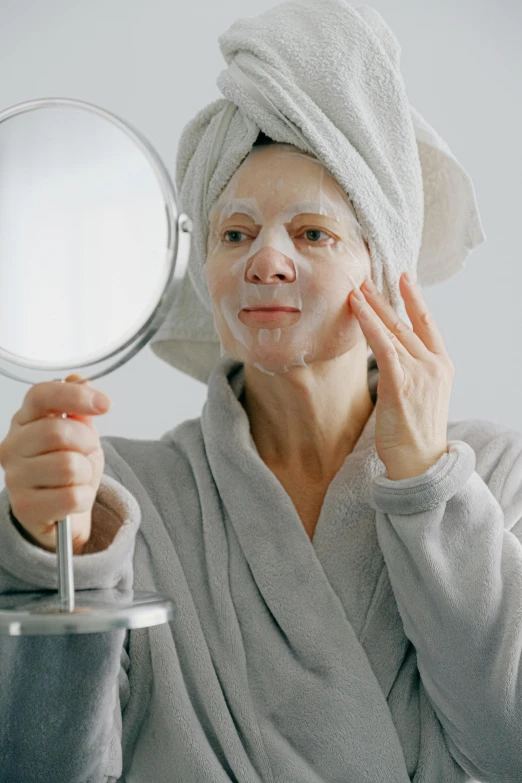 a woman with a towel on her head holding a mirror, wearing facemask, aging, wearing a grey robe, porcelain skin tone