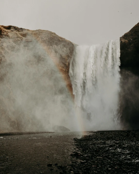 a person standing in front of a waterfall, by Terese Nielsen, pexels contest winner, renaissance, grey skies with two rainbows, thumbnail, dusty rock in background, glaciers