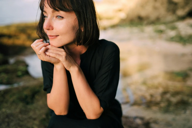 a woman sitting on a rock near a body of water, a portrait, inspired by Zoë Mozert, pexels contest winner, hurufiyya, sitting with wrists together, bjork smiling, slightly minimal, black