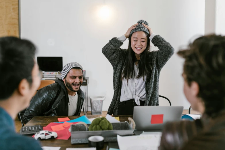 a group of people sitting around a wooden table, by Jang Seung-eop, trending on unsplash, happening, wearing a silly hat, in an office, beanie, she is laughing