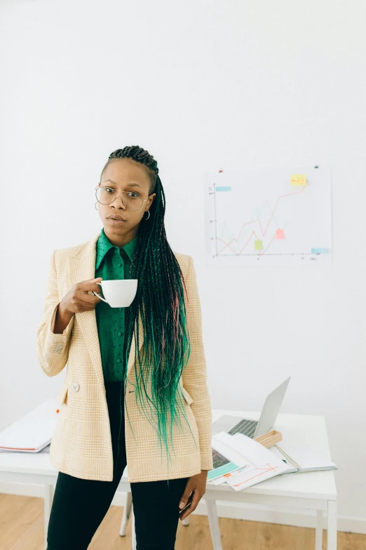 a woman standing in front of a desk holding a cup of coffee, by Dulah Marie Evans, trending on unsplash, afrofuturism, wearing green suit, concerned expression, ai researcher, non binary model