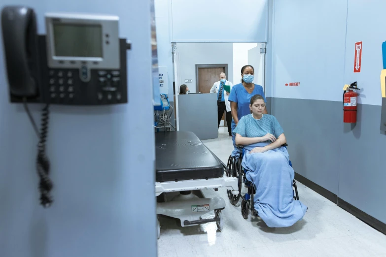 a man in a wheelchair in a hospital hallway, by Meredith Dillman, pexels, hurufiyya, surgical gown and scrubs on, aperture science test chamber, new mexico, 30 years old woman