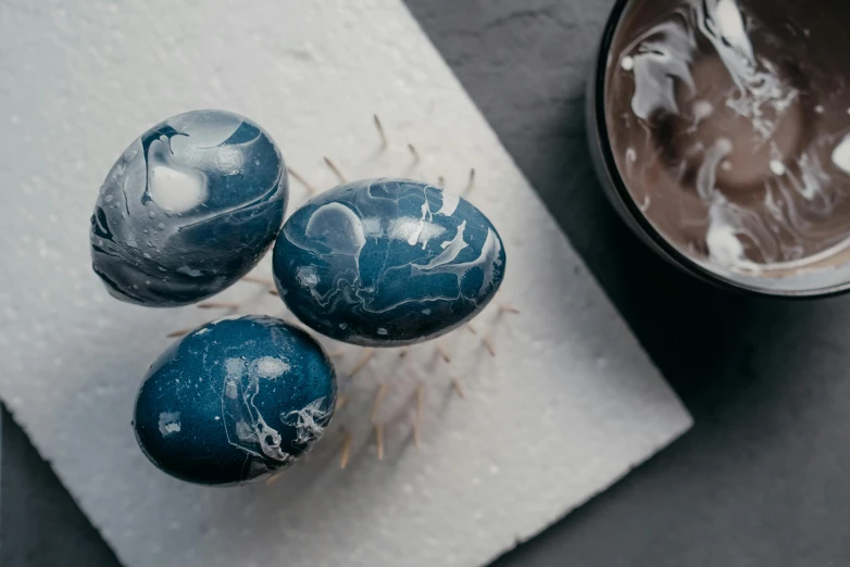 three blue marbled eggs sitting next to a cup of coffee, a marble sculpture, inspired by Hokusai, unsplash, process art, spikes on the body, hair like dark clouds, close-up product photo, monochrome