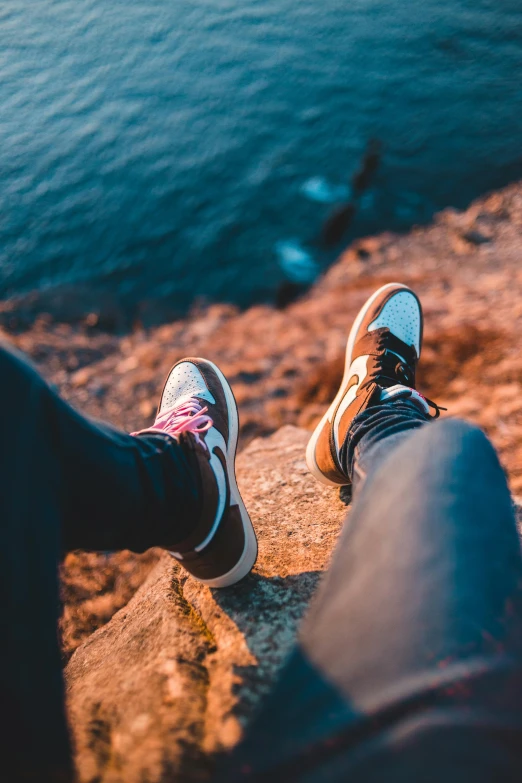 a person sitting on top of a rock next to a body of water, trending on pexels, realism, sneaker design, leg high, air shot, flirting