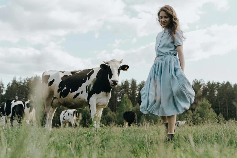 a woman in a blue dress standing in a field with cows, by Emma Andijewska, pexels contest winner, sadie sink, 15081959 21121991 01012000 4k, flowing milk, greta thunberg