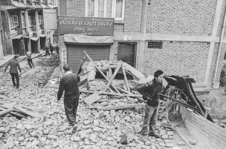 a couple of men standing next to a pile of rubble, flickr, nepali architecture buildings, on sidewalk, 1 9 7 5 photo, debris flying around