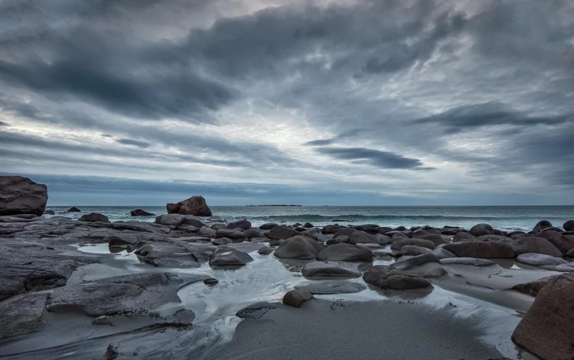 a large body of water sitting on top of a sandy beach, a portrait, with lots of dark grey rocks, skies, ominous evening, new zealand