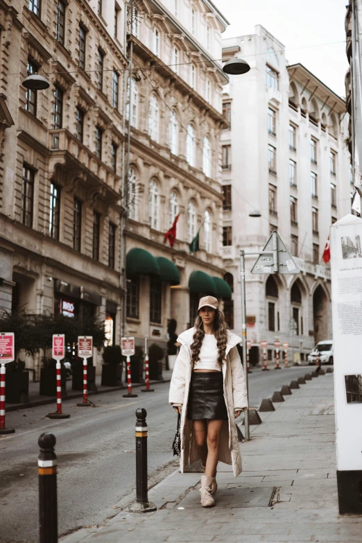 a woman walking down a street past tall buildings, trending on pexels, short skirt and a long jacket, budapest, wearing a chocker and cute hat, posing for a picture