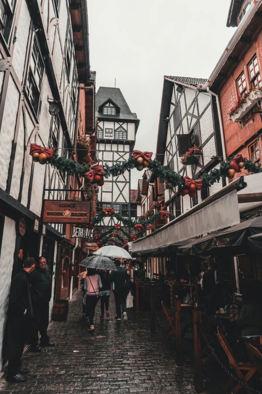 people walking down a cobblestone street under an umbrella, a photo, unsplash contest winner, art nouveau, timbered house with bricks, holiday, decorations, red building