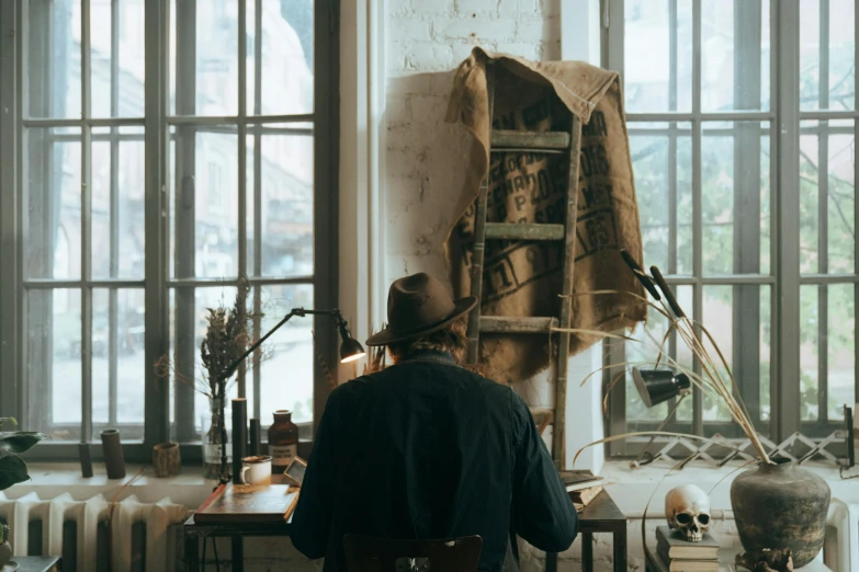 a man sitting at a desk in front of a window, inspired by Raymond Leech, pexels contest winner, wearing a straw hat and overalls, there is a skull over a table, long shot from the back, with a bunch of stuff