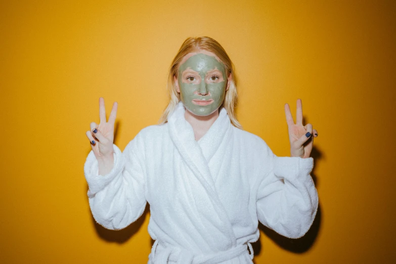 a woman in a bathrobe making a peace sign with her hands, inspired by Elsa Bleda, trending on pexels, green facemask, mud on face, a blond, gold mask