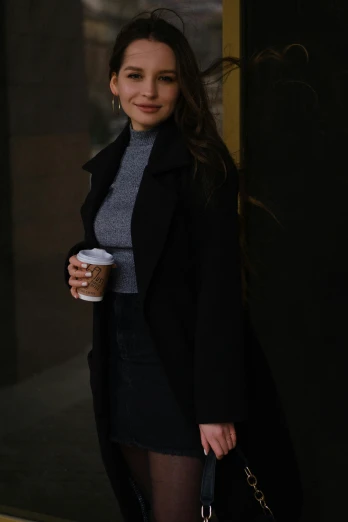 a woman standing outside of a building holding a cup of coffee, by Robbie Trevino, black coat, portrait sophie mudd, f / 2 0, gif