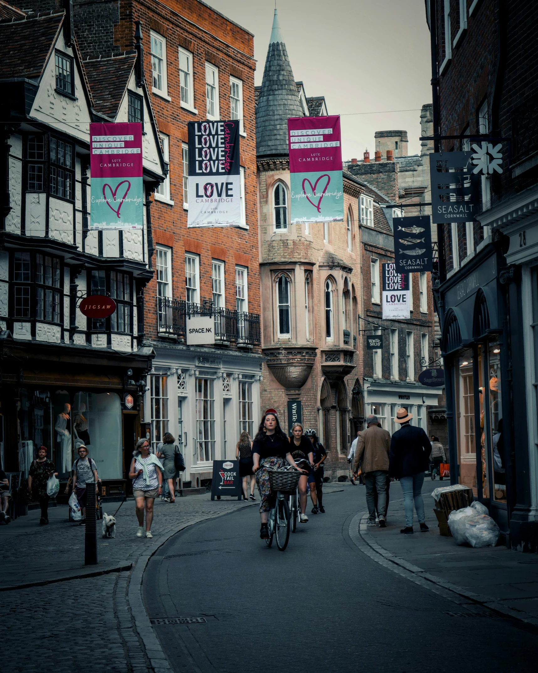 a group of people riding bikes down a street, poster art, by IAN SPRIGGS, pexels contest winner, river stour in canterbury, cloth banners, people walking in street, photograph of the city street