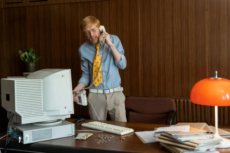 a man standing next to a desk talking on a phone, by Peter Churcher, looks like domhnall gleeson, old computers, gettyimages, 15081959 21121991 01012000 4k