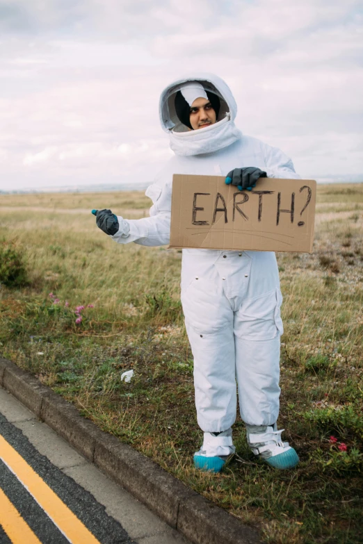 a person in a space suit holding a sign, unsplash, happening, earthy, standing in road, full costume, earthwave