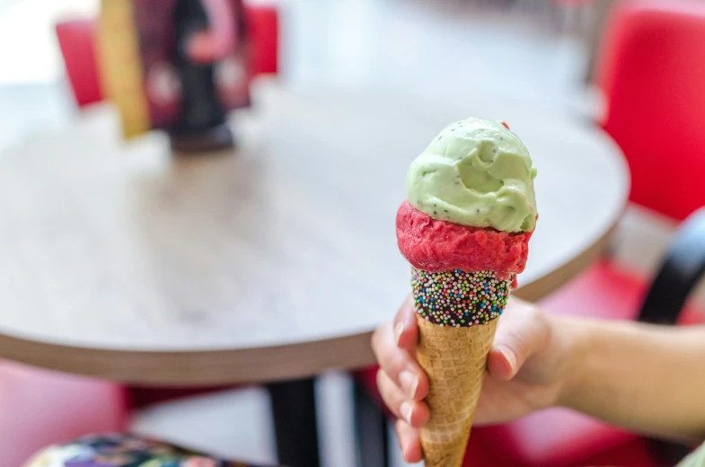 a person holding an ice cream cone with sprinkles, green bright red, hand on table, subtle detailing, coloured photo