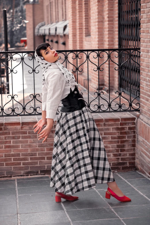 a woman standing in front of a brick building, an album cover, inspired by Modest Urgell, unsplash, arabesque, plaid skirt, looks like audrey hepburn, at a fashion shoot, white and black clothing