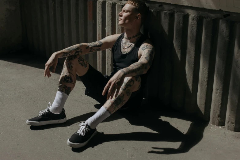 a man with tattoos sitting against a wall, inspired by Seb McKinnon, pexels contest winner, bauhaus, wearing : tanktop, high soles, non binary model, harsh sunlight