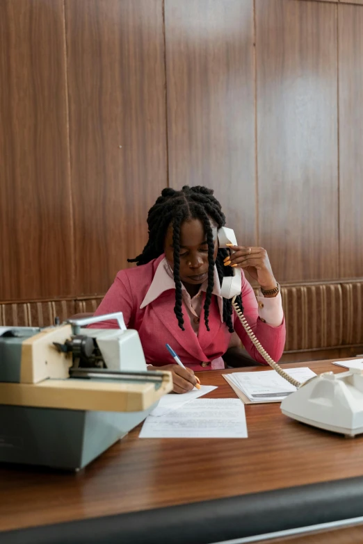 a woman sitting at a desk talking on a phone, young thug, government archive, brown and pink color scheme, taken in 2 0 2 0