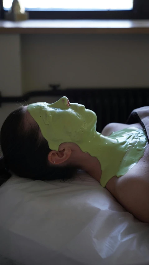 a woman laying on top of a bed next to a window, inspired by Elsa Bleda, reddit, green facemask, made of glowing wax and ceramic, candy treatments, profile image