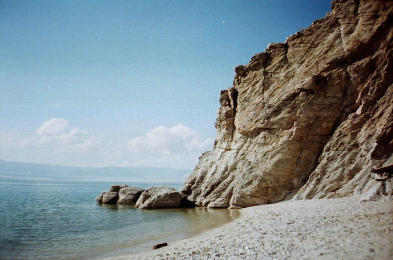 a man standing on top of a sandy beach next to the ocean, a colorized photo, unsplash, romanticism, chalk cliffs above, ((rocks)), orthodox, 1990s photograph