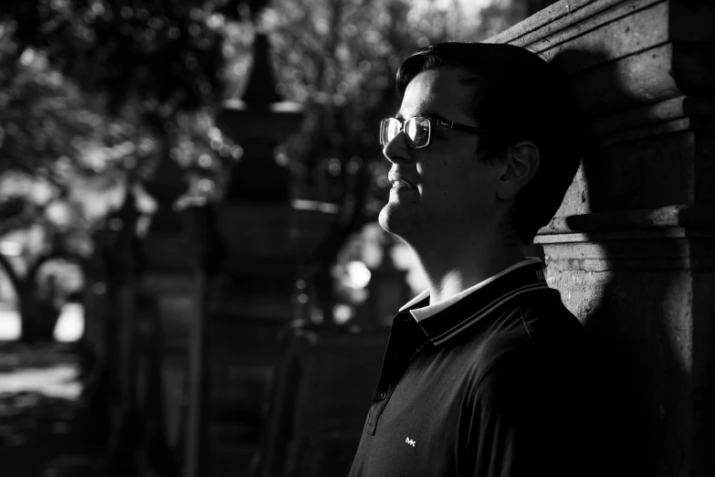 a black and white photo of a man leaning against a wall, by Jan Rustem, brandon sanderson, backlighting, in a city park, man with glasses