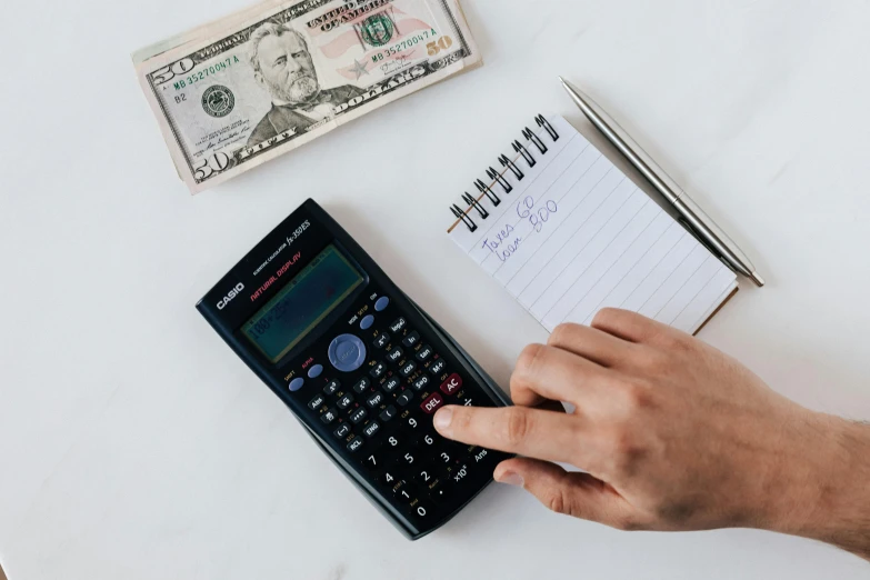 a person using a calculator next to a stack of money, a photo, by Carey Morris, pexels, 🦩🪐🐞👩🏻🦳, erak note, ad image, realistic »