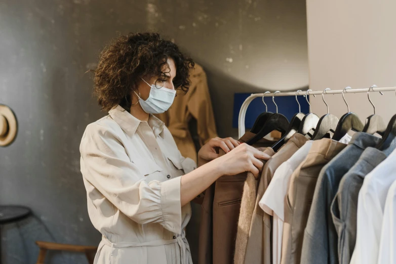 a woman standing in front of a rack of clothes, trending on pexels, happening, wearing facemask, wearing a brown, professional picture, caretaker